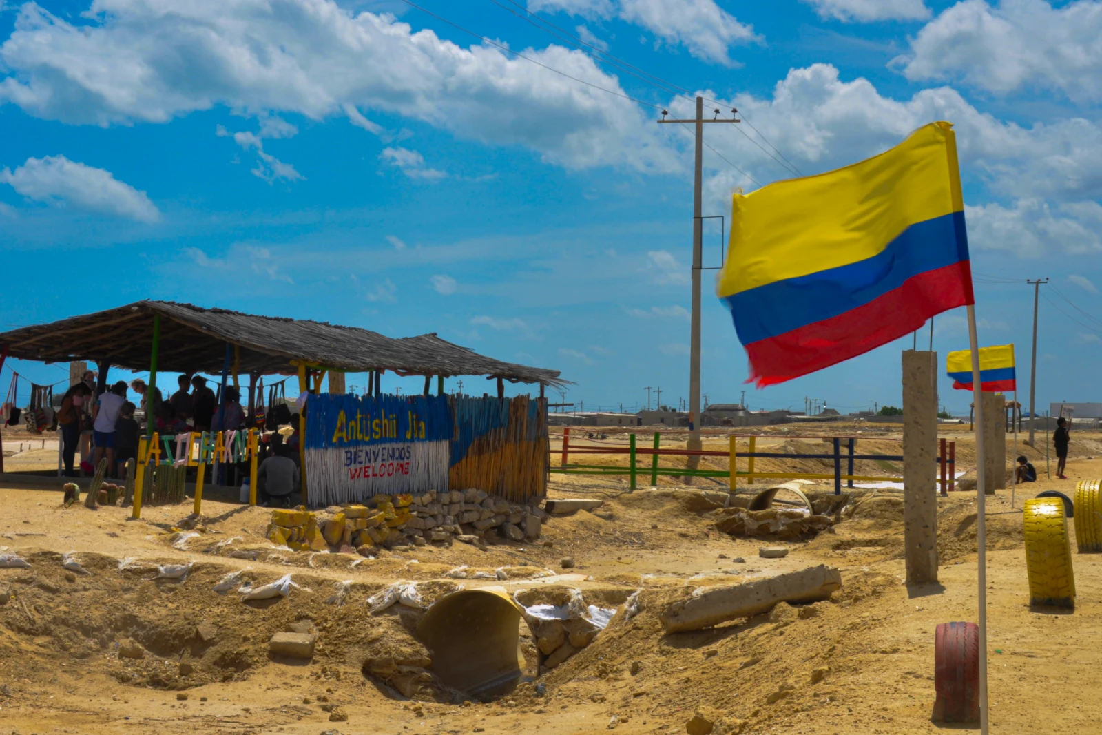 Drapeau colombien sur un mât près d'un étal au toit de palmes, sur une halte en terre battue, avec des poteaux électriques et des nuages blancs.