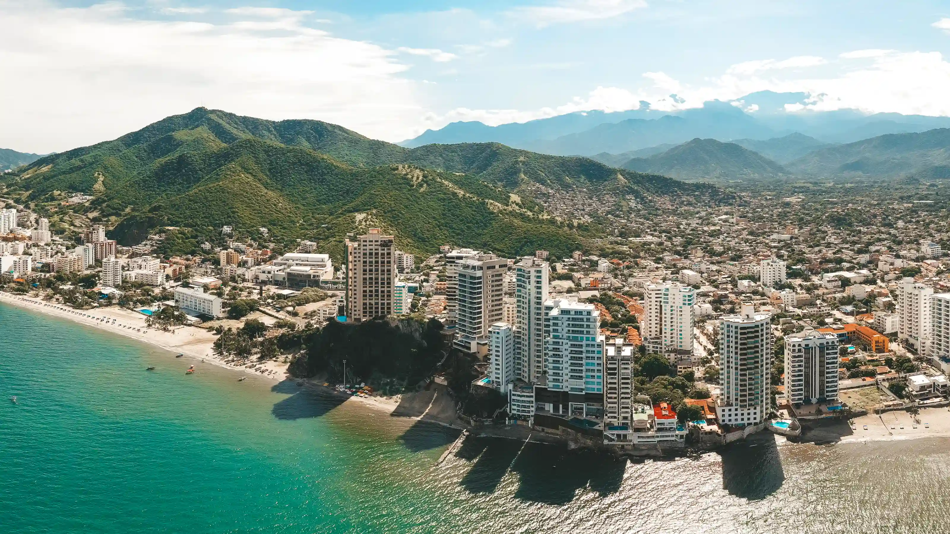 Vue aérienne d'immeubles face à une plage turquoise à Santa Marta, avec des quartiers à flanc de colline et des montagnes vertes au fond.