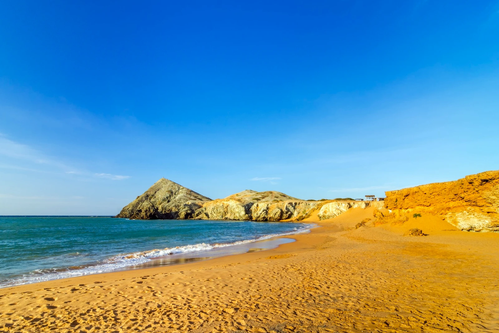 Plage de sable ocre avec le Pilón de Azúcar et d'autres collines au fond, mer bleue aux vagues douces, à Cabo de la Vela.