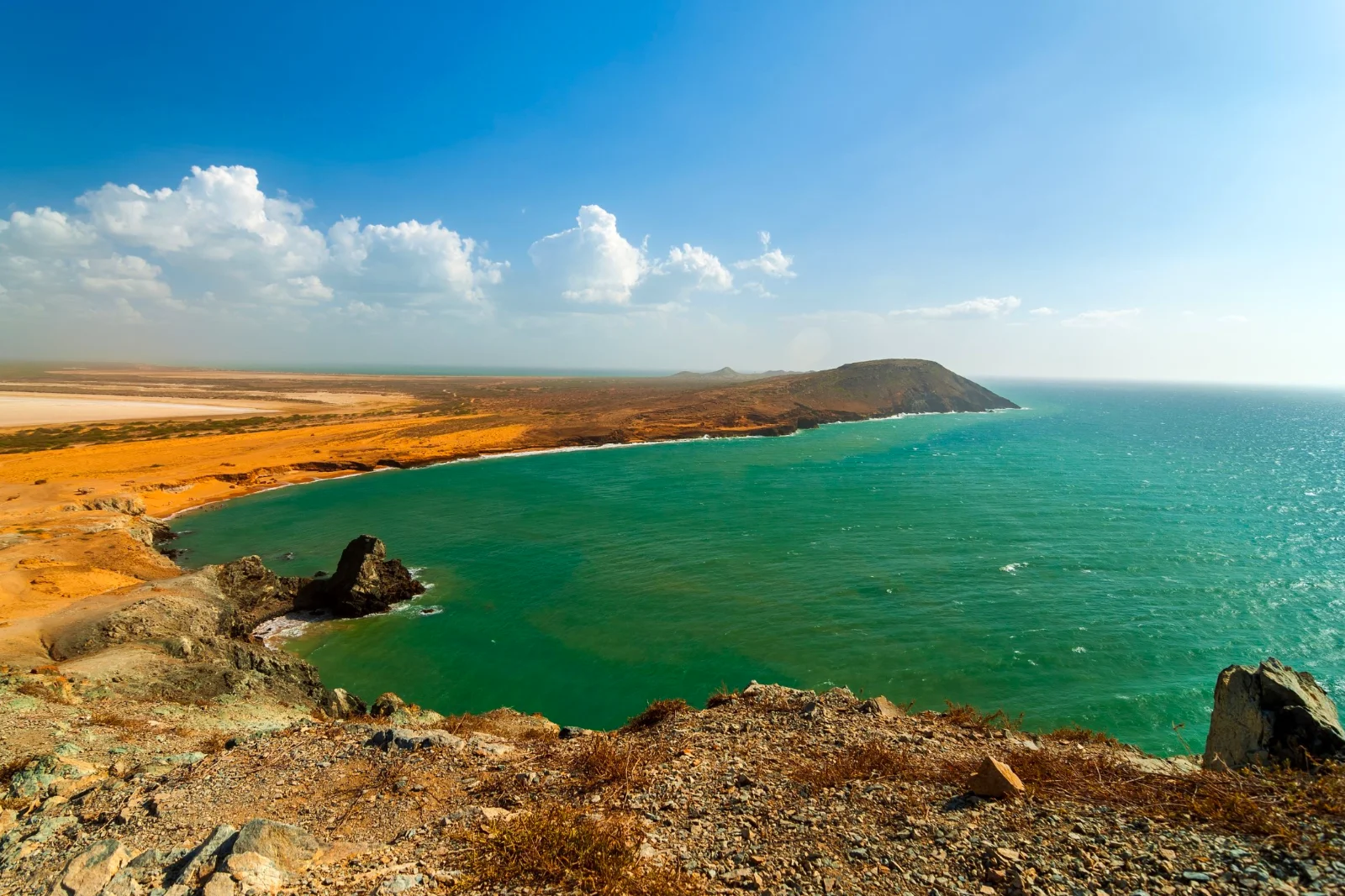 Baie d'eau vert émeraude vue depuis une falaise, avec une côte de sable ocre, des rochers épars et un cap au fond sous des nuages blancs.