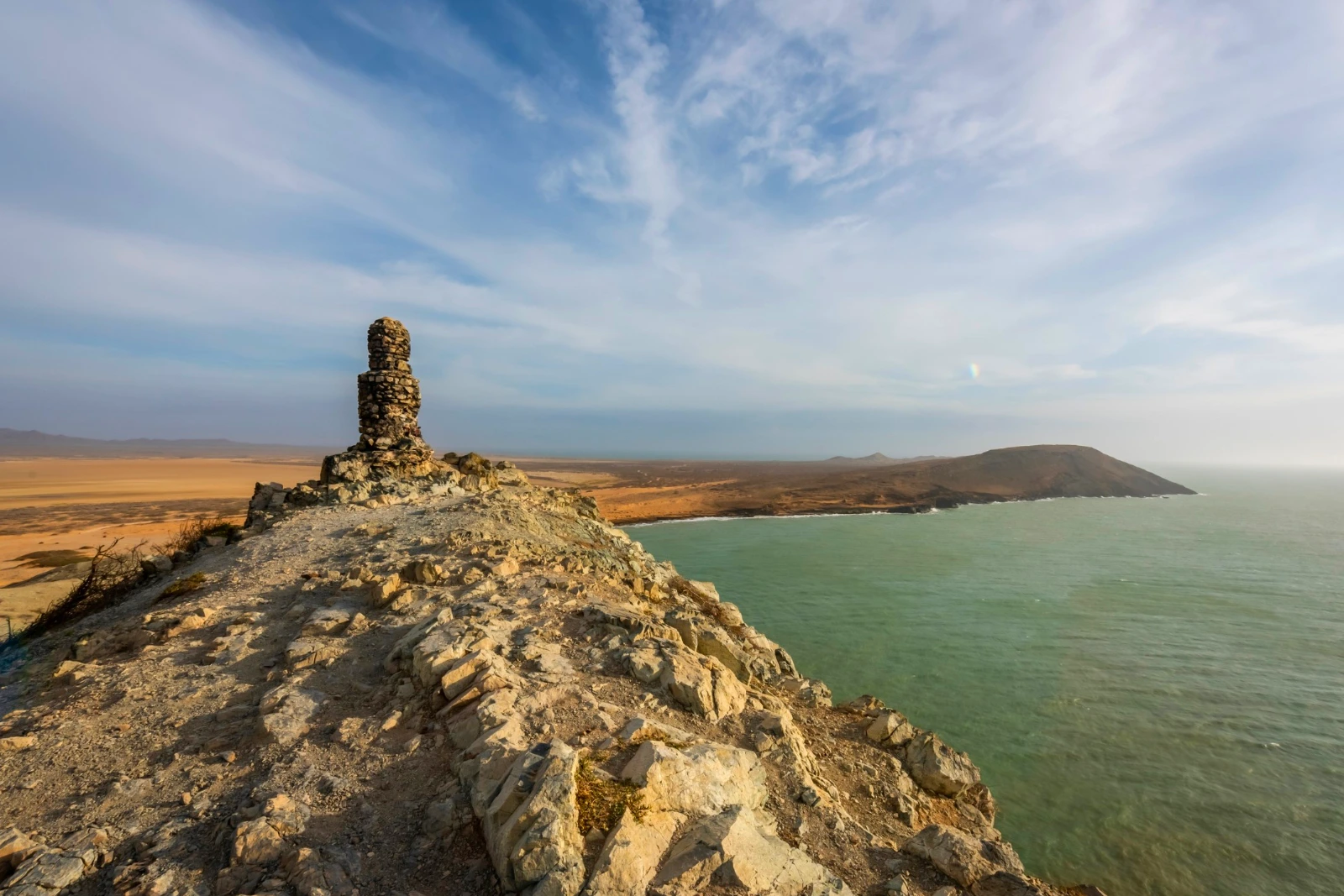 Empilement de pierres sur une pointe rocheuse face à une baie verte et un cap, sous des nuages d'altitude et la lumière du soir.