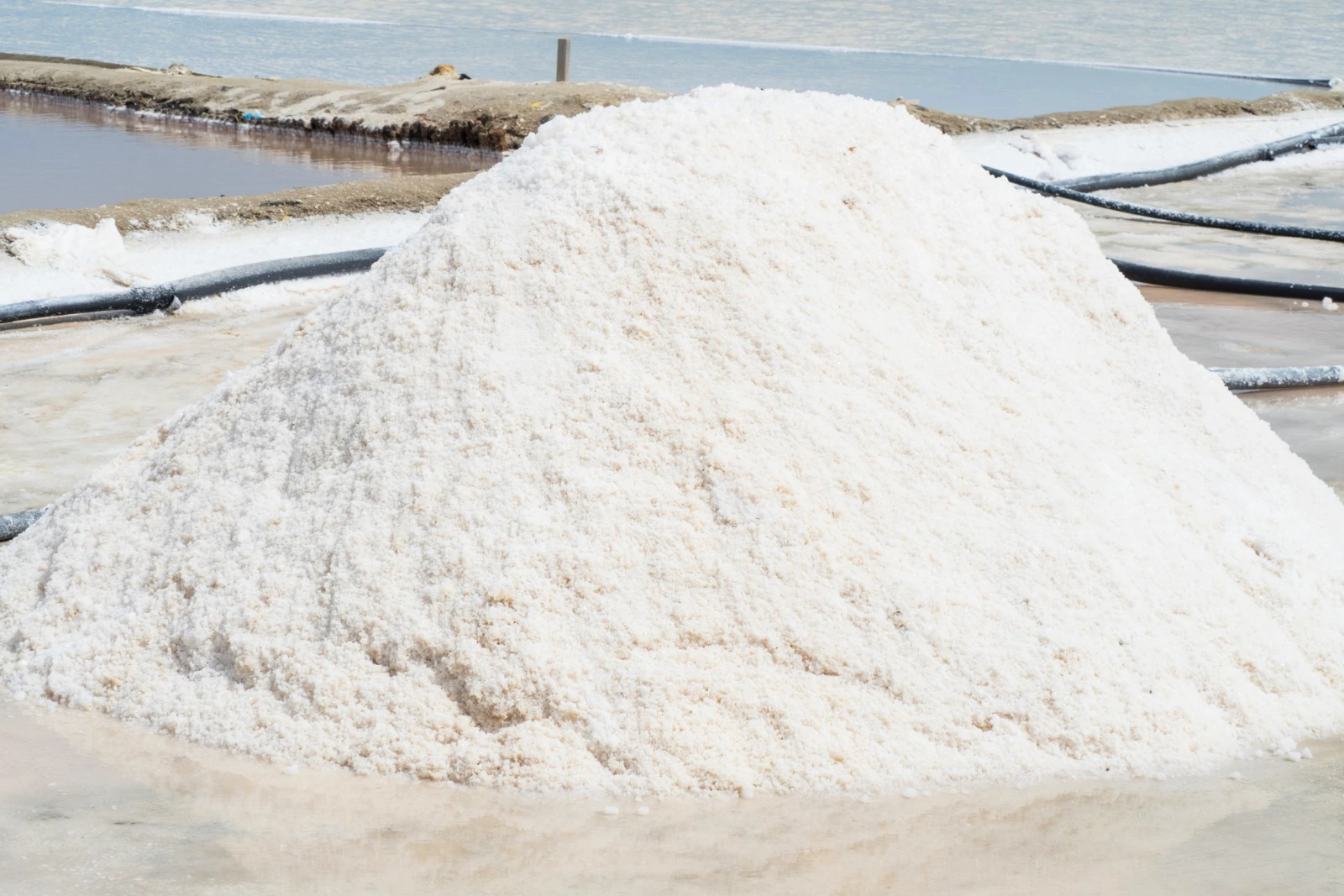 Monticule de sel blanc près de bassins d'évaporation et de tuyaux noirs, dans des salines sous un ciel bleu pâle.