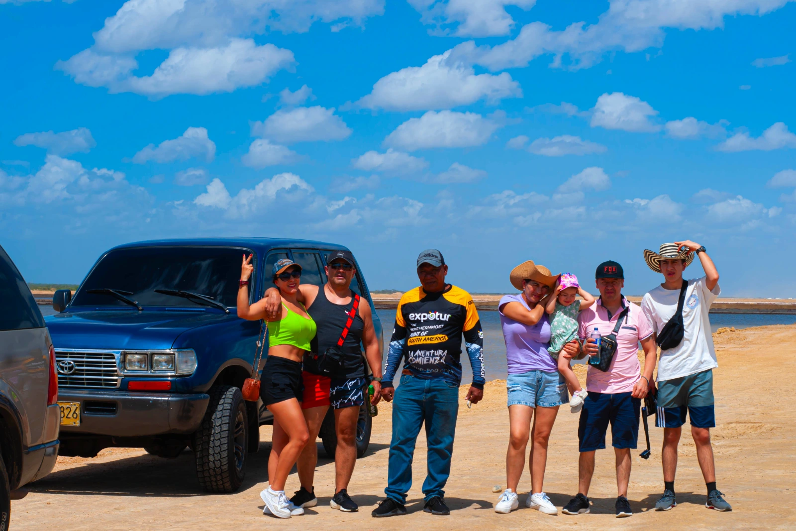 Un groupe de voyageurs et un guide posent près d'un 4×4 bleu sur une étendue de sable, avec de l'eau et un ciel bleu nuageux.