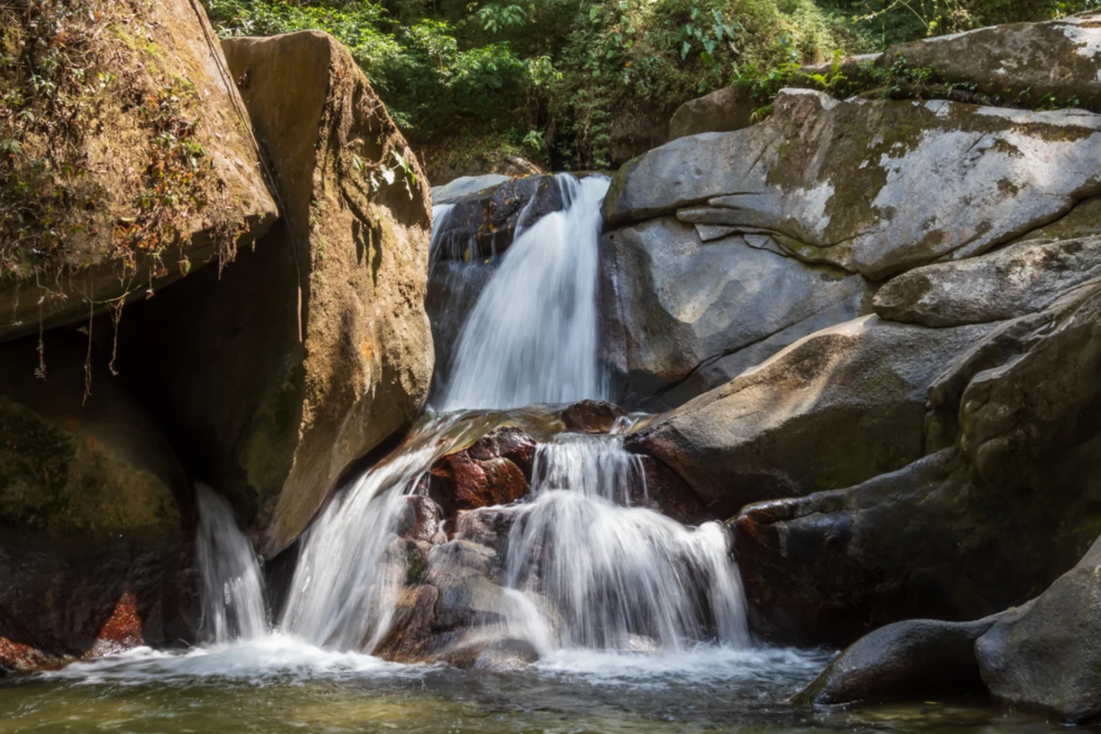 Cascada que baja en varios saltos entre rocas grandes y lisas hasta un pozo de agua quieta, rodeada de selva, en Minca.