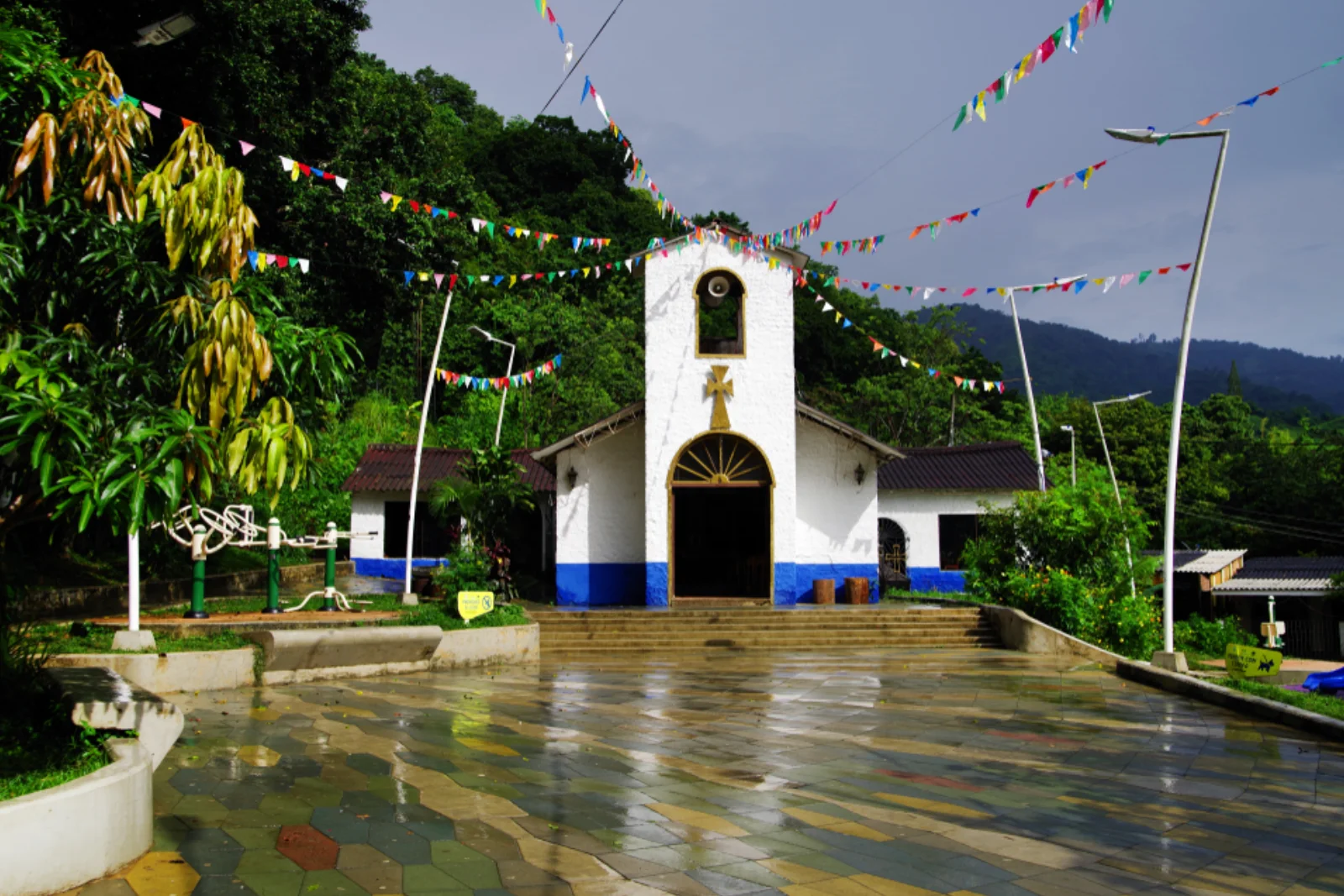 Iglesia blanca de Minca con zócalo azul y banderines de colores sobre la plaza mojada por la lluvia, con cerros verdes y cielo gris.