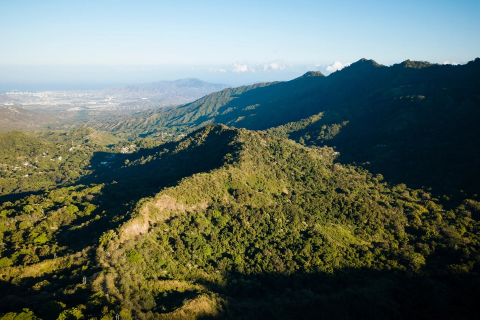 Cordilleras cubiertas de bosque con luz lateral de la tarde y, a lo lejos, la costa del mar Caribe con una ciudad bajo el cielo despejado.