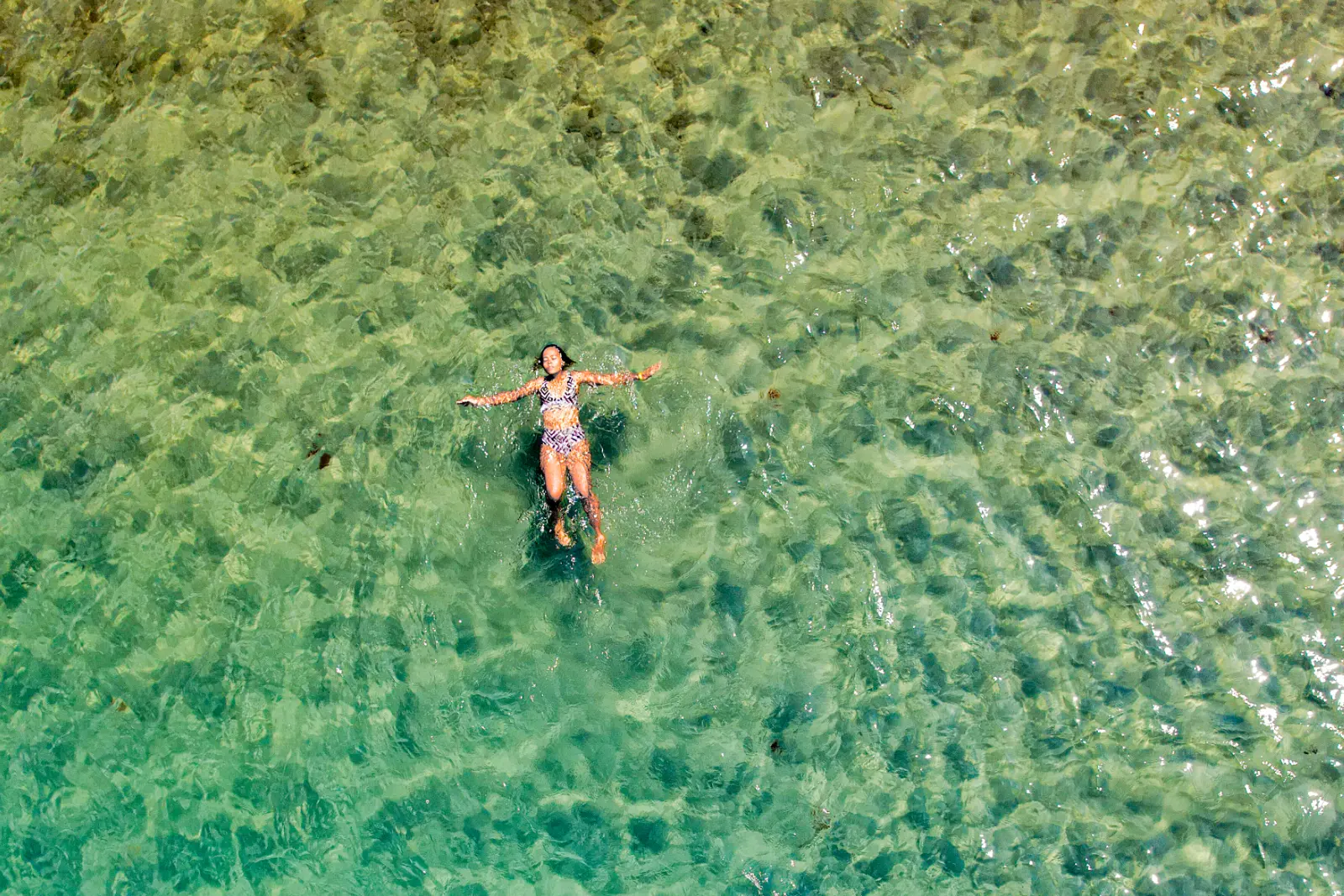 Vue zénithale d'une femme qui flotte sur le dos, bras écartés, dans une eau verte et transparente au-dessus d'un fond de sable.