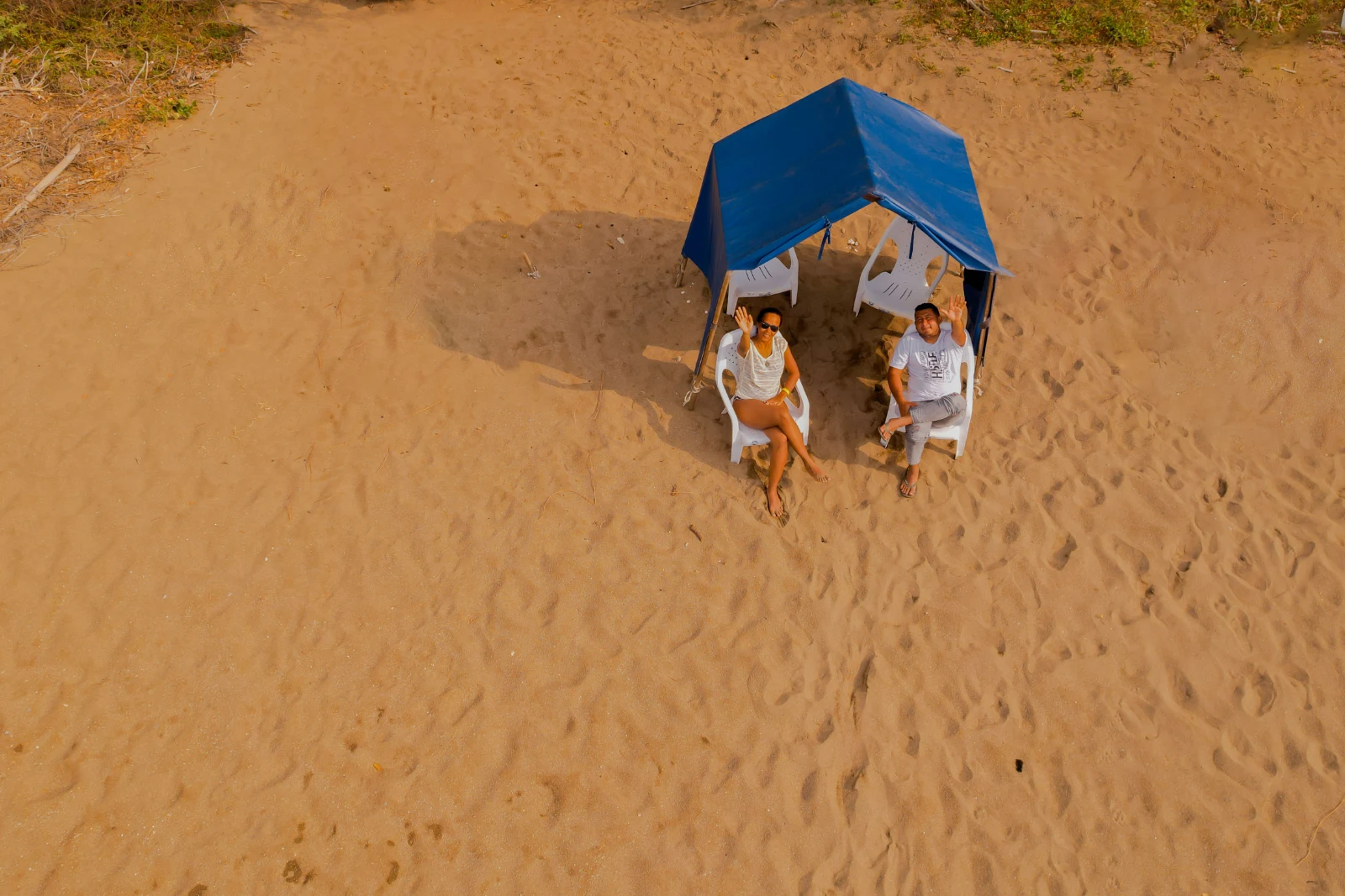Vue zénithale d'un couple assis sur des chaises blanches sous un auvent bleu sur le sable, saluant de la main vers le haut.