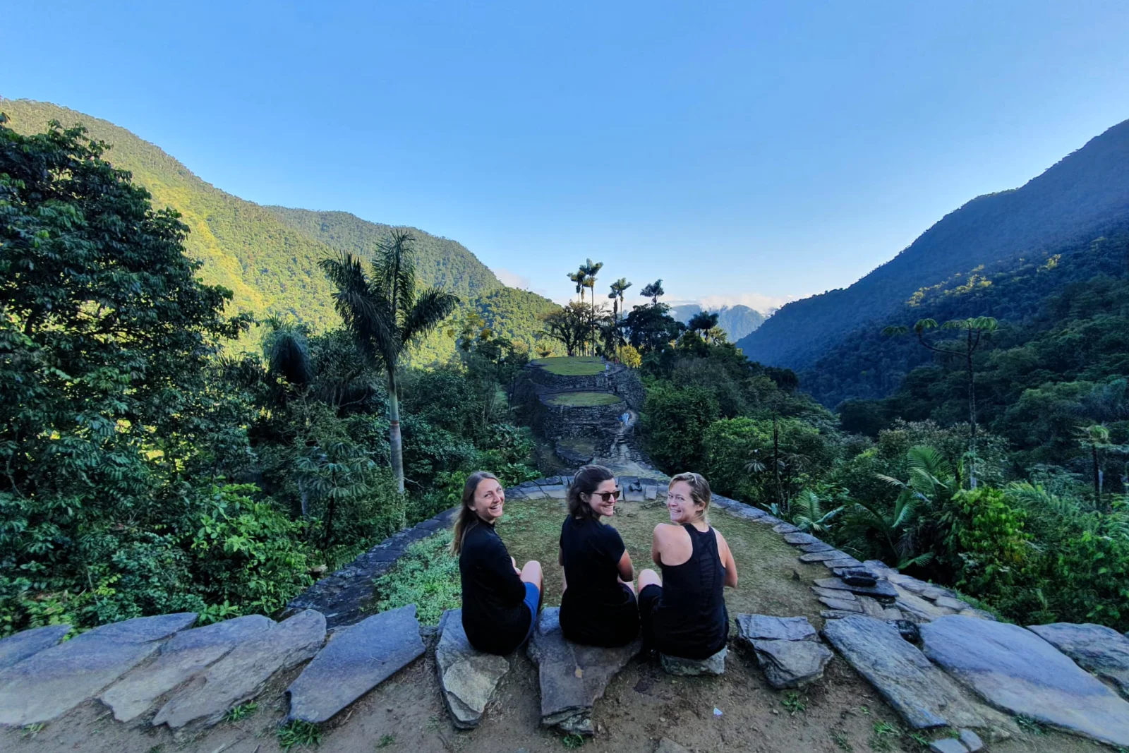 Three hikers sitting on the edge of a stone terrace look back at the camera, with terraces, palms and Sierra Nevada mountains behind.