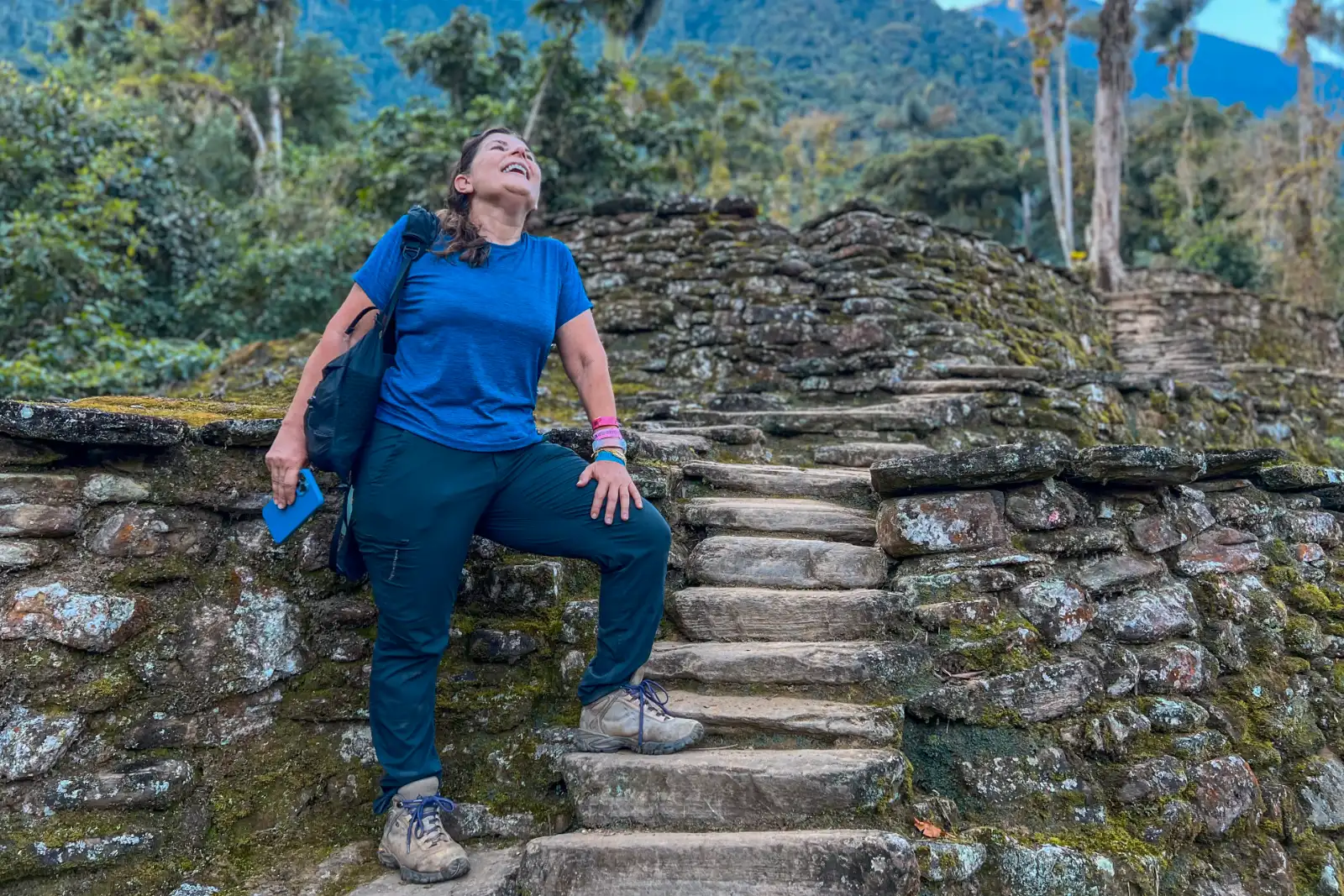 A hiker in a blue T-shirt poses, smiling, on a stone stairway of the Ciudad Perdida (Teyuna) terraces, with palms and forest behind.