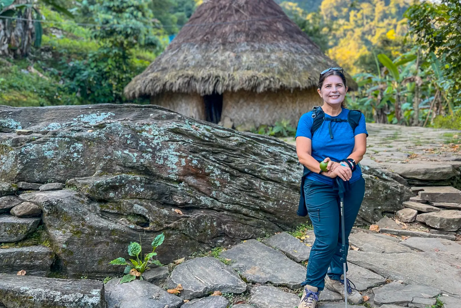 A hiker in a blue T-shirt with a trekking pole smiles in front of a palm-thatched hut and a large boulder, on a flagstone floor.