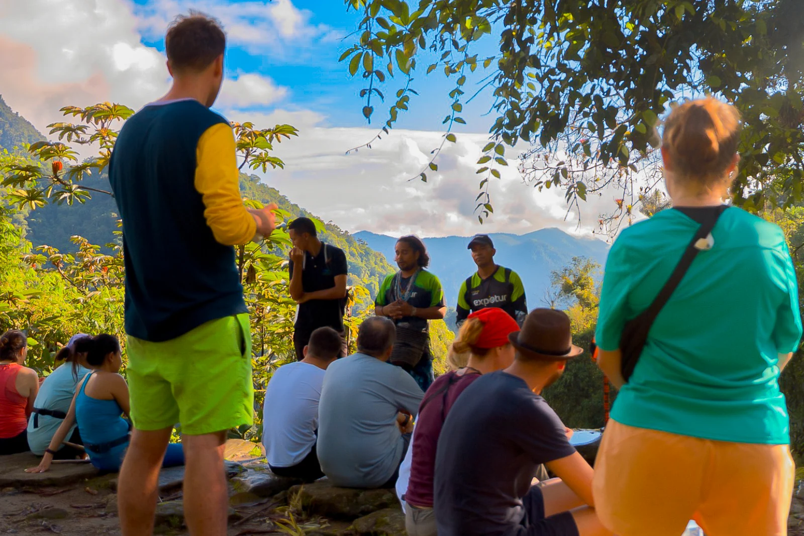 A group of hikers sitting on a terrace listens to two guides in green shirts, with Sierra Nevada mountains and clouds behind.