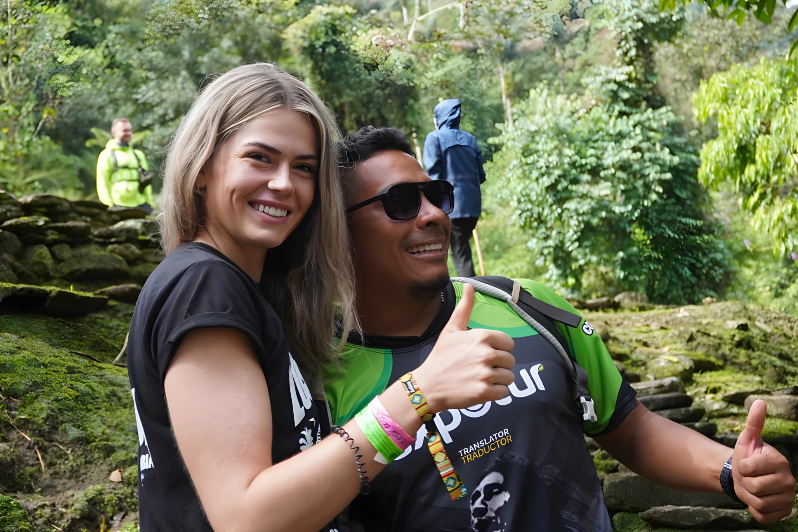 A blond traveler and a guide in an Expotur shirt pose close up with a thumbs-up among moss-covered stone walls.