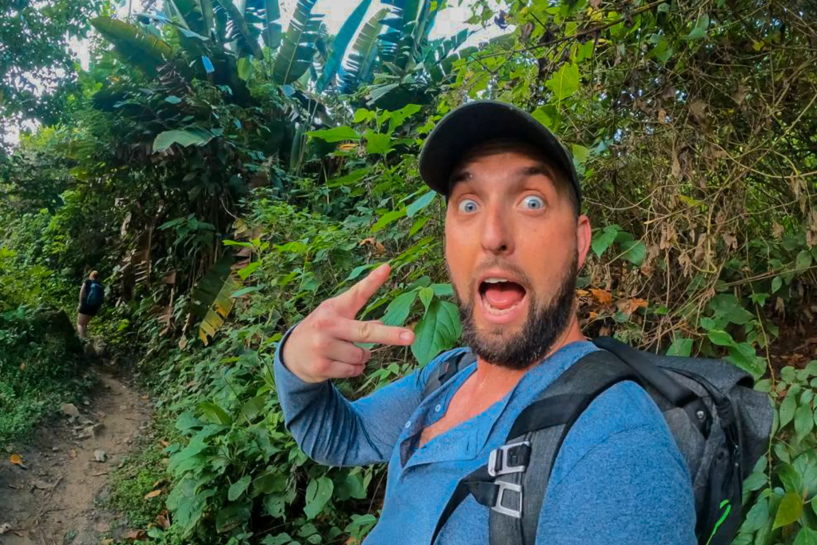 Selfie of a hiker in a cap and backpack making a surprised face on a narrow forest trail.