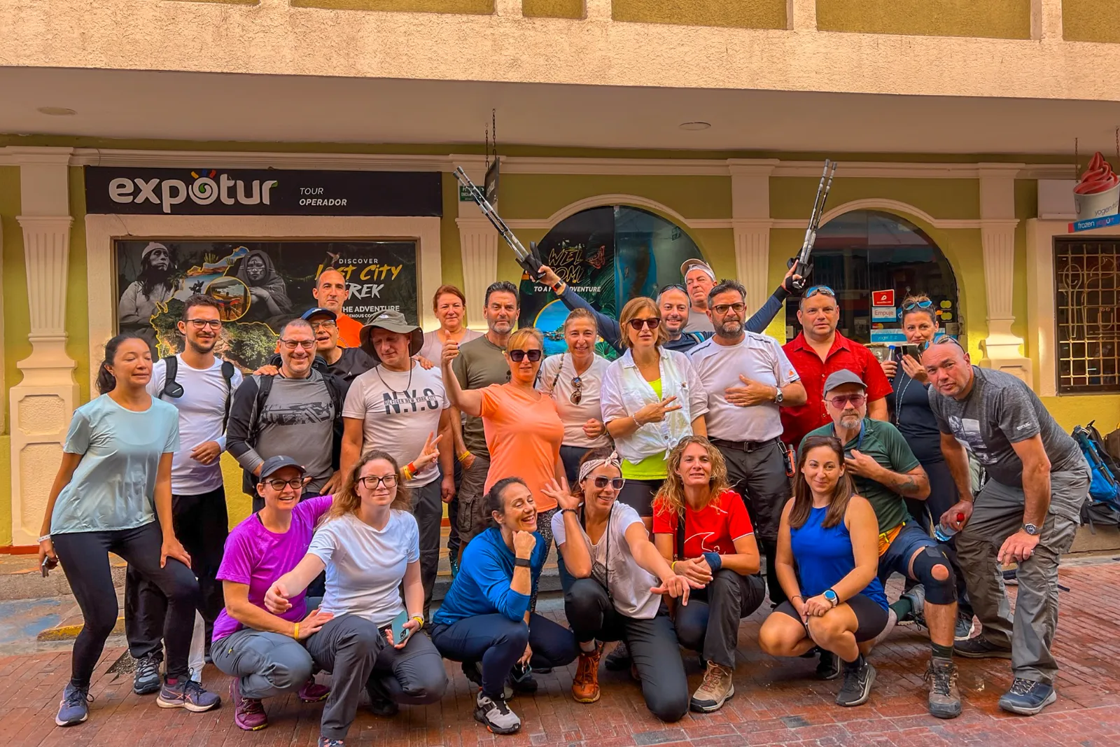 A large group of hikers poses on the street in front of an office with an Expotur sign and a Lost City Trek poster.