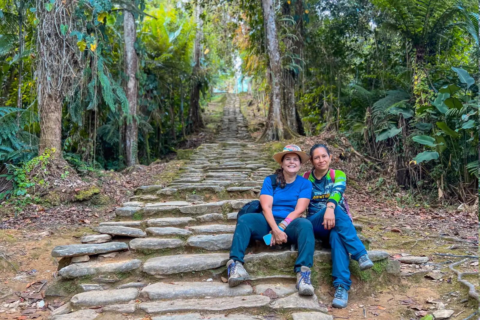 Deux femmes, l'une coiffée d'un chapeau et l'autre en maillot de guide Expotur, assises souriantes sur un long escalier de pierre en forêt.