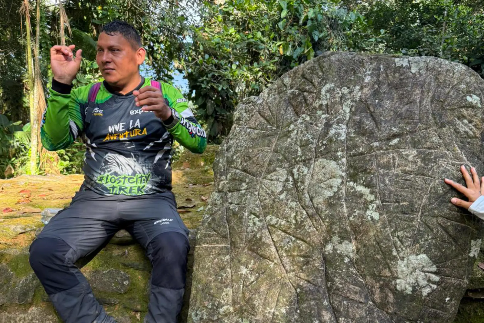 Un guide en t-shirt vert assis près d'un gros rocher aux lignes gravées explique avec les mains, dans la forêt de la Sierra Nevada.