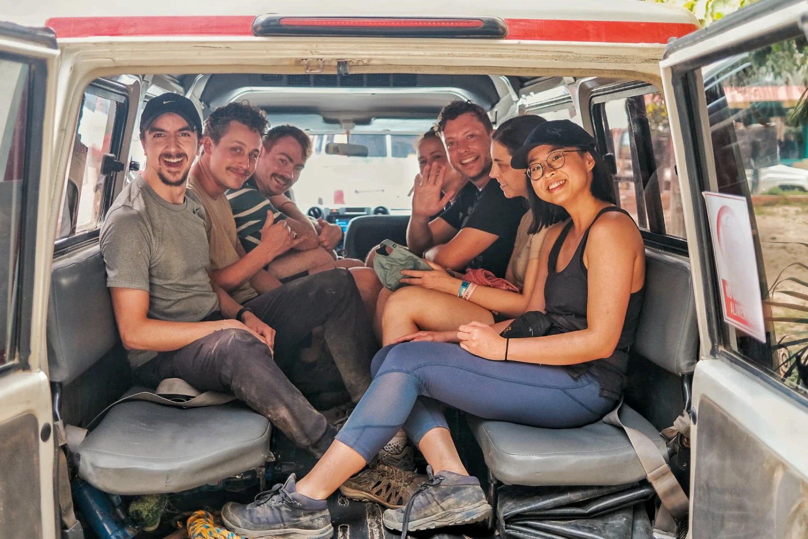 Sept jeunes voyageurs assis à l'arrière d'un 4×4 sourient et saluent l'objectif par la porte ouverte.
