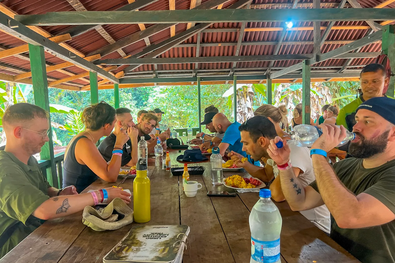 Randonneurs autour d'une longue table en bois sous un toit de tôle dans un camp, avec des assiettes de fruits et des bouteilles d'eau.