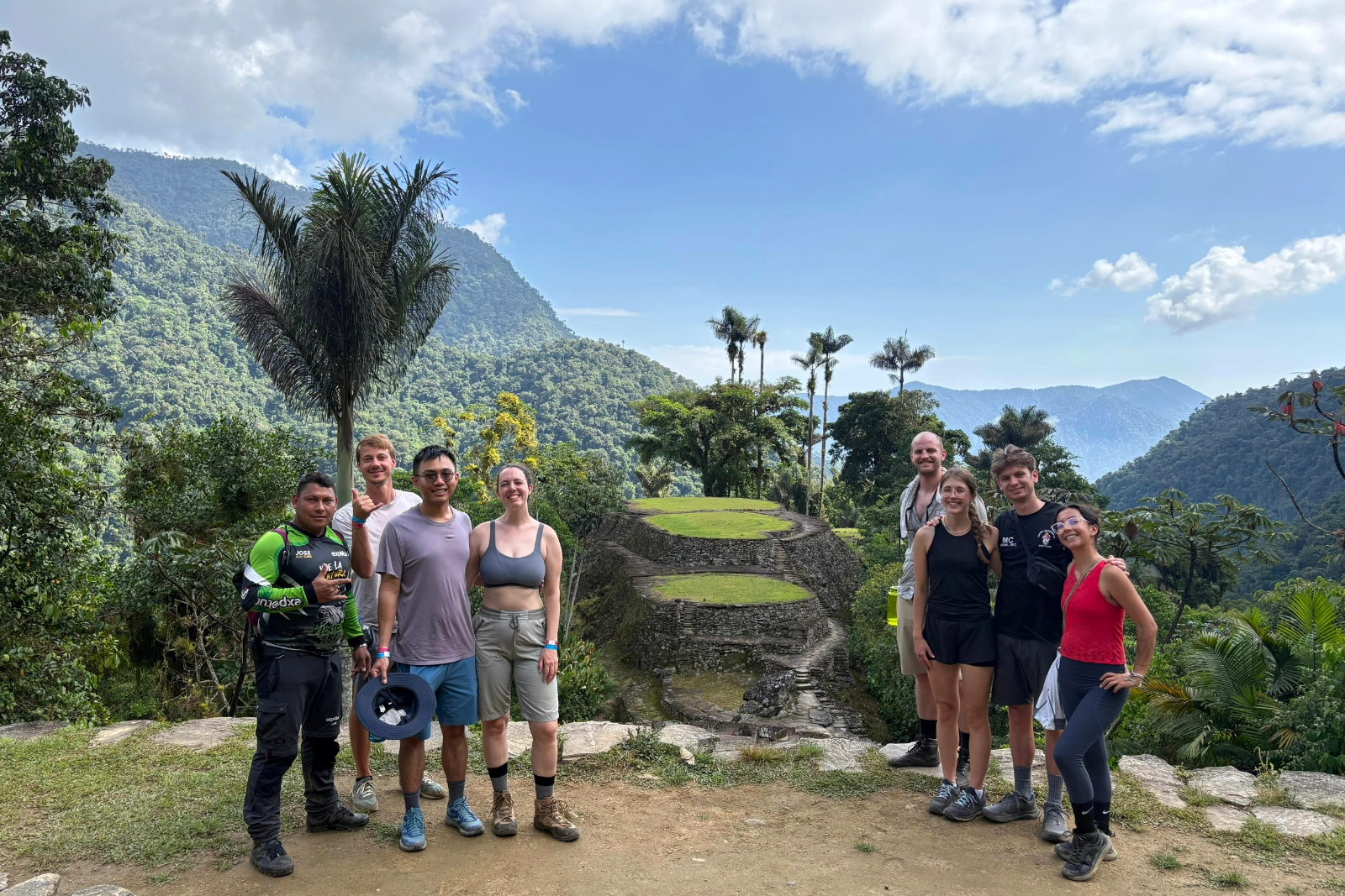 Un groupe de randonneurs et un guide en t-shirt vert posent devant les terrasses circulaires de Ciudad Perdida (Teyuna), palmiers et montagnes.