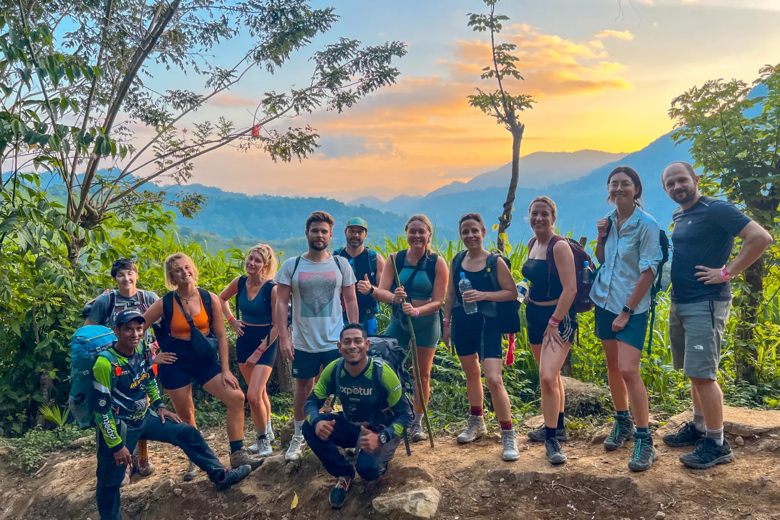 A group of hikers and guides poses on a trail at sunset, with Sierra Nevada mountains and an orange sky behind them.