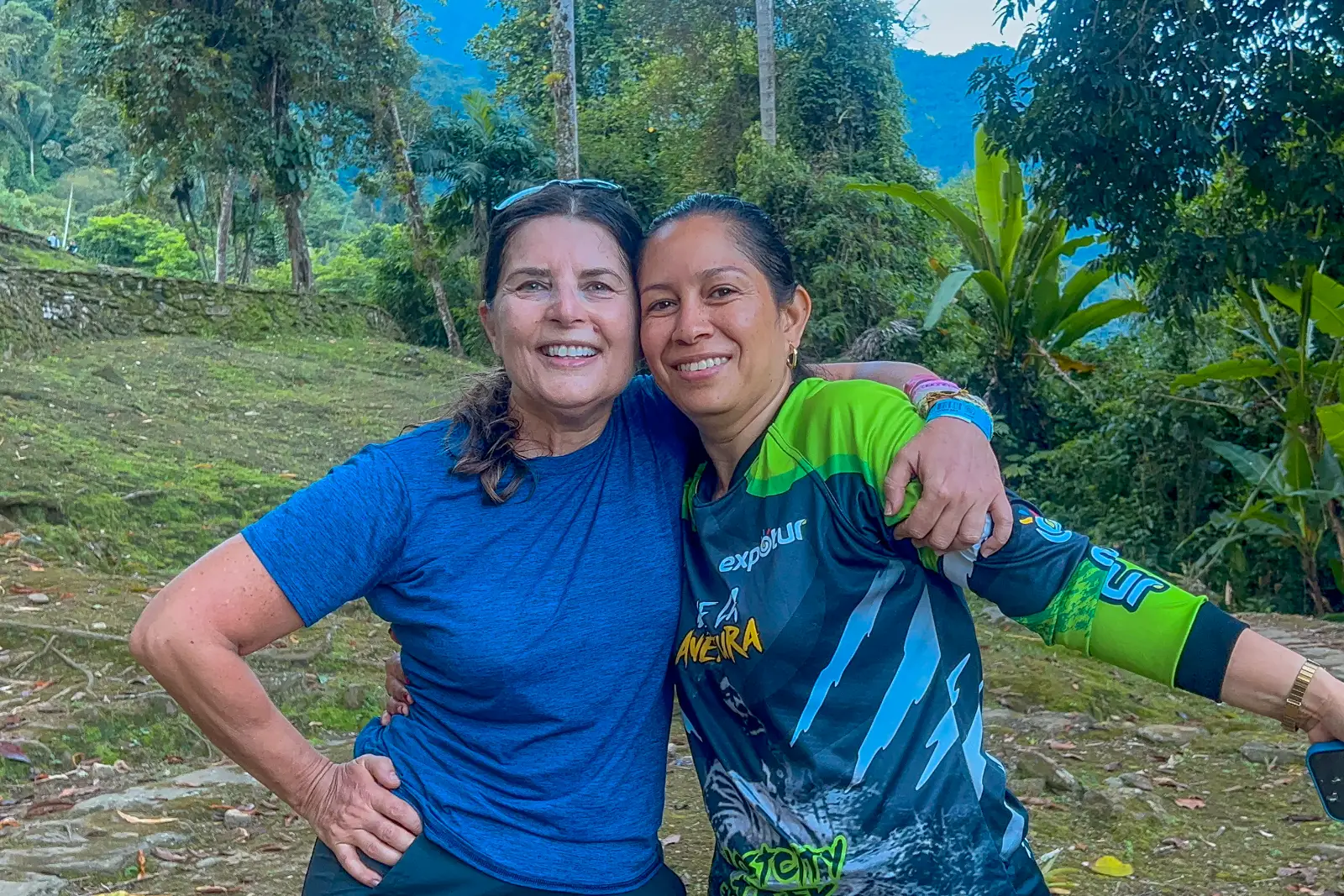 Two women smile arm in arm, one in a blue T-shirt and one in an Expotur guide shirt, on a grassy terrace with trees behind.