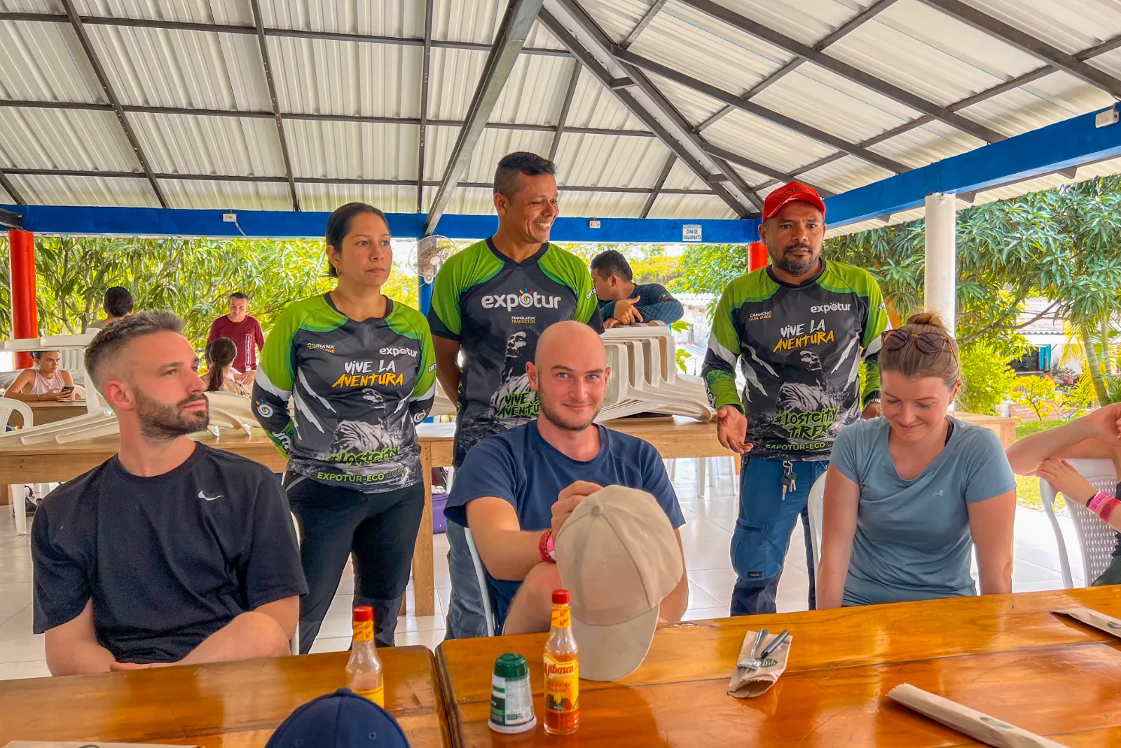 Travelers and guides in Expotur shirts around a wooden table under a tin roof, in an open-air dining area.