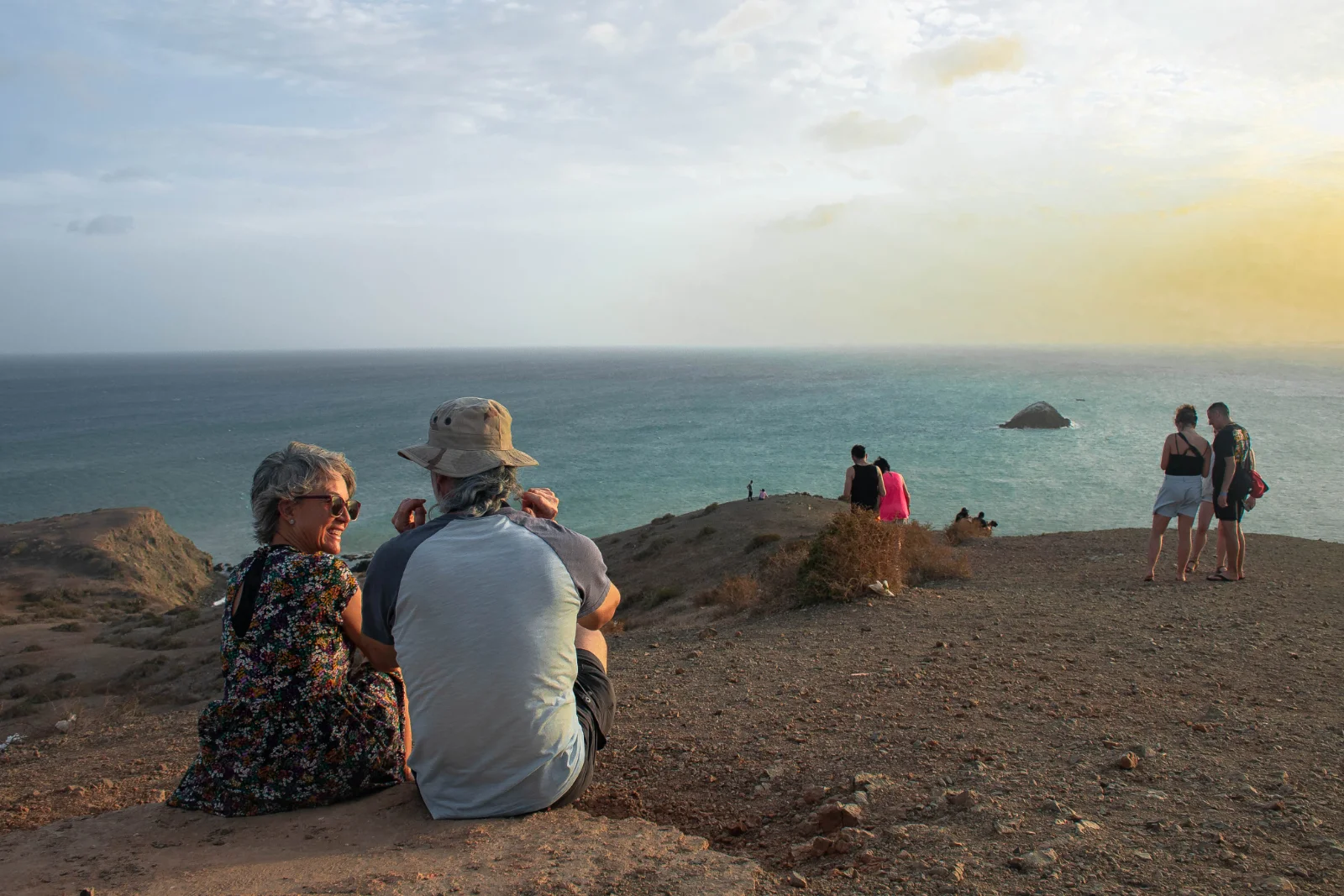 Visiteurs assis et debout sur un promontoire côtier face à la mer, avec un îlot rocheux au loin et le ciel voilé de l'après-midi.