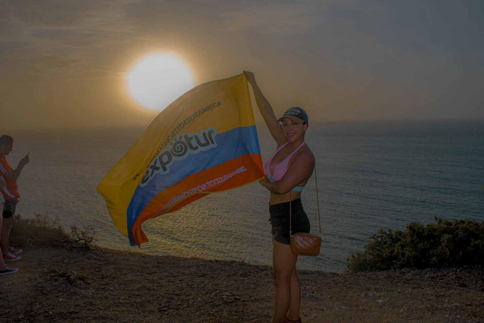 Une voyageuse à contre-jour brandit un grand drapeau au logo d'Expotur sur une falaise face à la mer, le soleil bas derrière elle.