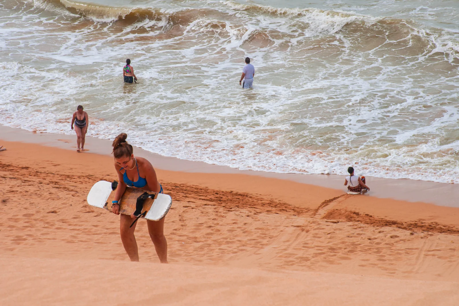 Une voyageuse gravit une dune au bord de la mer avec une planche pour glisser sur le sable, tandis que d'autres se baignent dans les vagues.