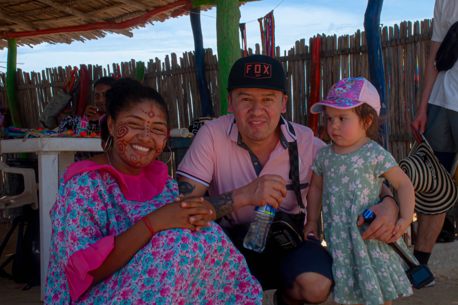Femme wayuu au visage peint et en manta imprimée, assise près d'un voyageur et d'une fillette sous une enramada où pendent des mochilas.