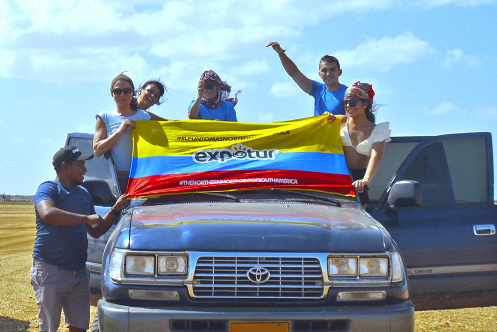 Un groupe de voyageurs tient un drapeau colombien au logo d'Expotur sur un 4×4 bleu, dans une plaine désertique sous un ciel bleu.
