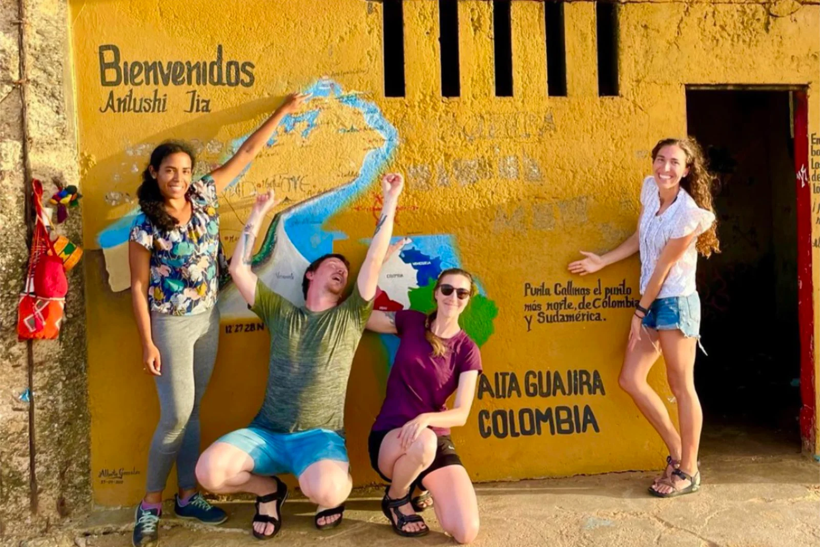 Quatre voyageurs posent devant un mur jaune peint d'une carte et des mots « Bienvenidos » et « Alta Guajira Colombia ».